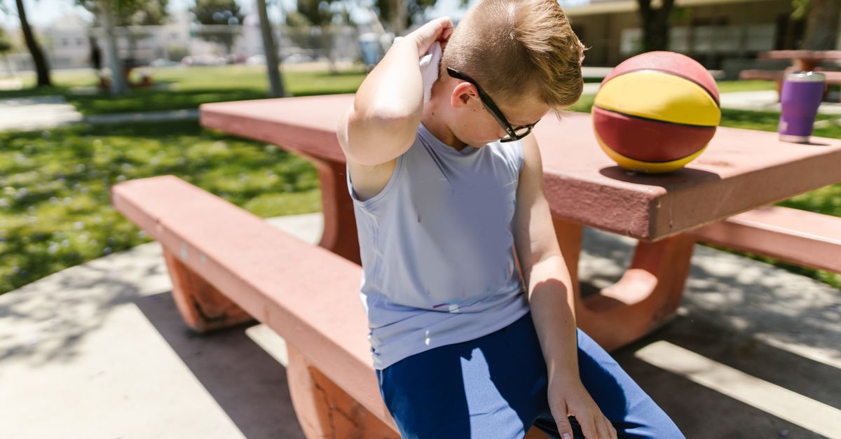 A young boy sitting on a bench at a park, resting with a basketball nearby.
